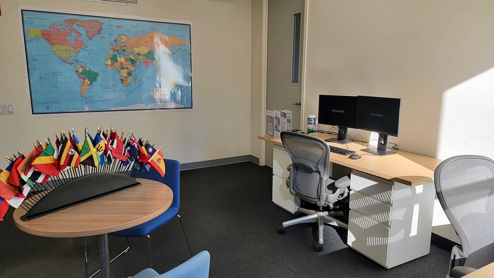 room with a world map on the wall, a desk with a computer, and a table with miniature world flags.