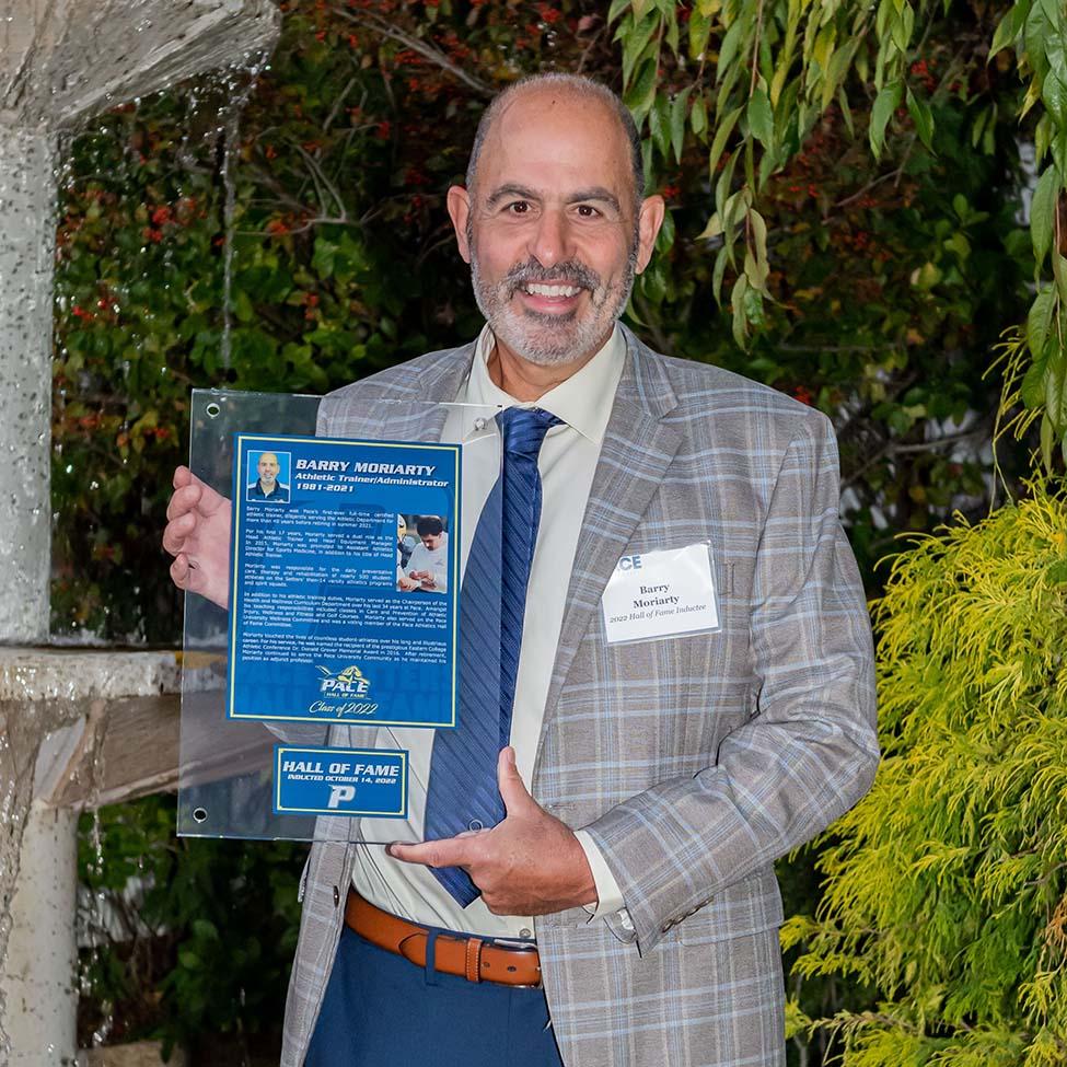 Barry Moriarty, wearing a grey suit jacket and blue tie, holds his Hall of Fame plaque