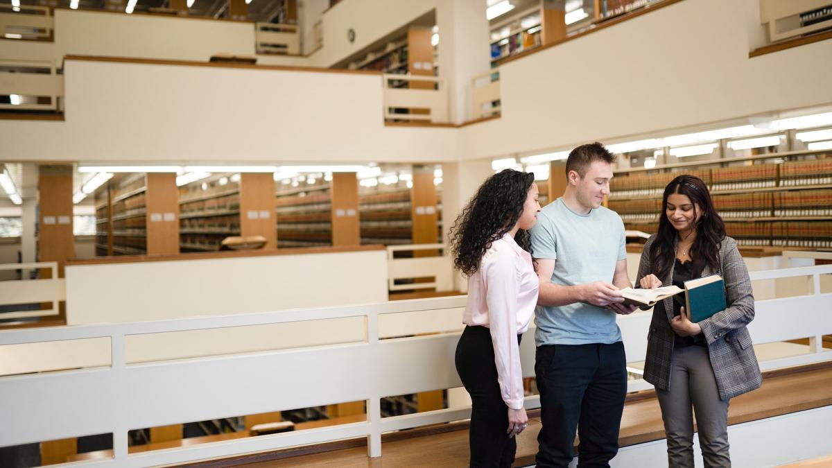 Group of students looking at a book, standing in the Elisabeth Haub School of Law library.