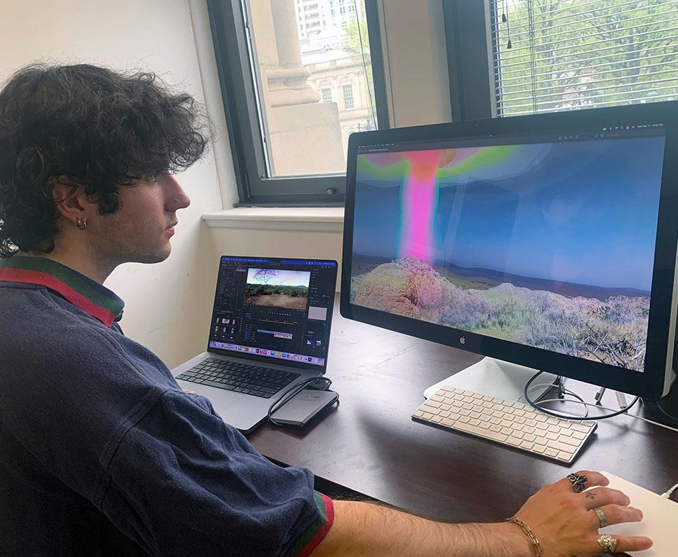 Pace University student Ben Pfiefer working on artwork on a desktop and laptop for Art professor Jillian McDonald's DUMBO Projection Project art exhibition