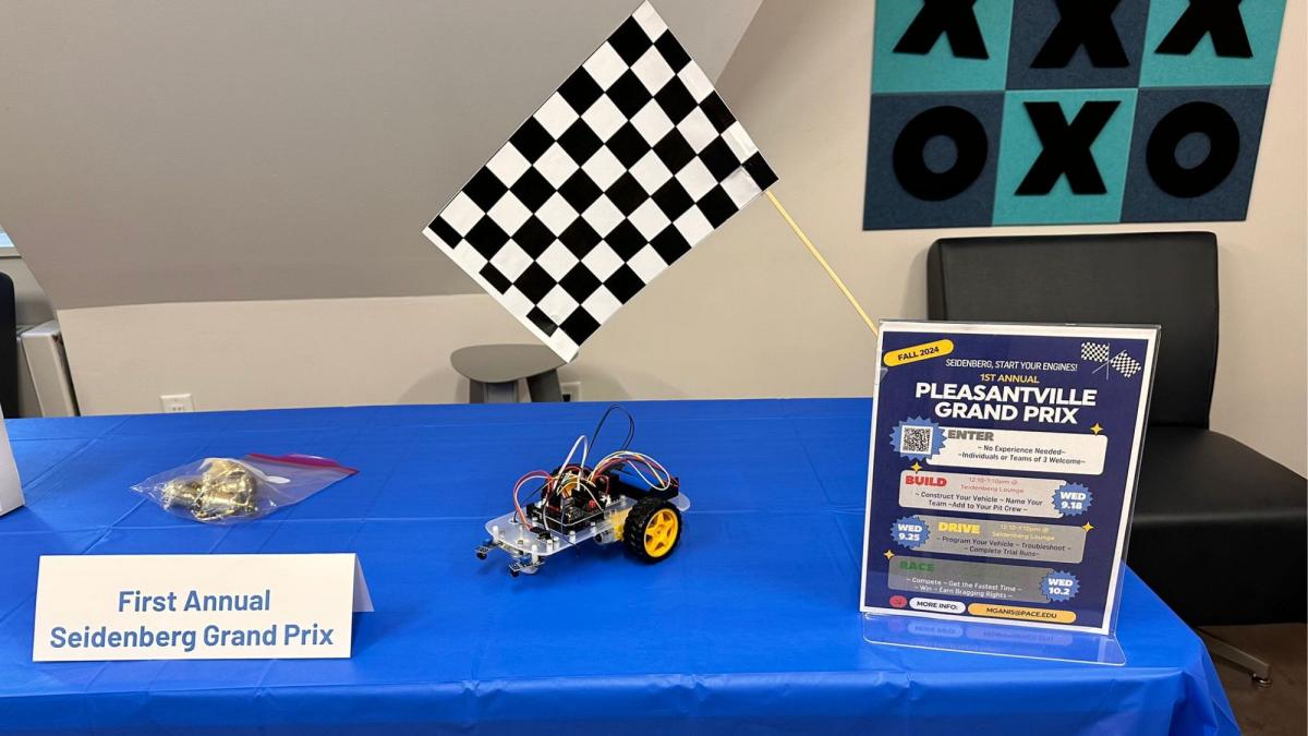 An informational table for the 1st Annual Pleasantville Grand Prix at Pace University. On the table are a flyer for the event, a checkered flag, and a small robot car built by students.