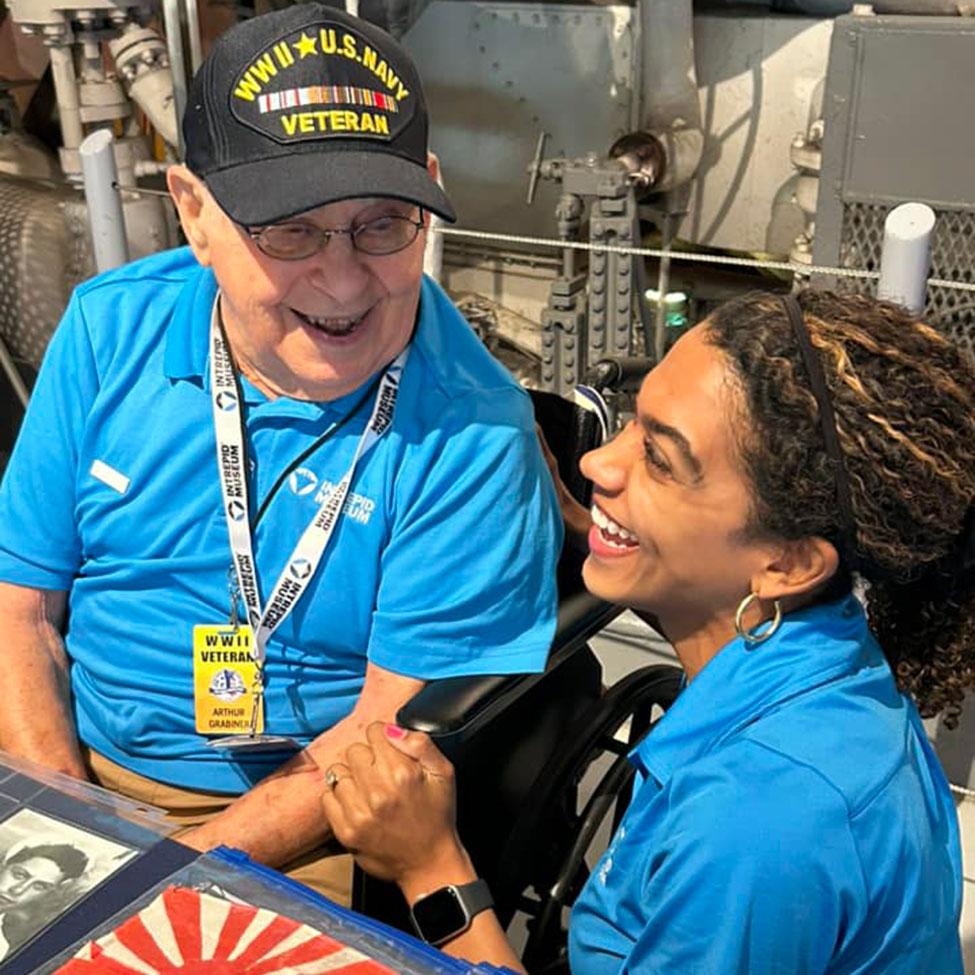 Pace University Alumni Arthur Grabiner '50 laughing with a young volunteer while appearing to speak with visitors at the Intrepid Museum.