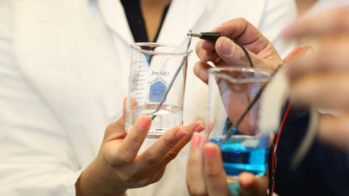 Student and professor in lab coats connecting a blue liquid with a clear one in beakers by a wire