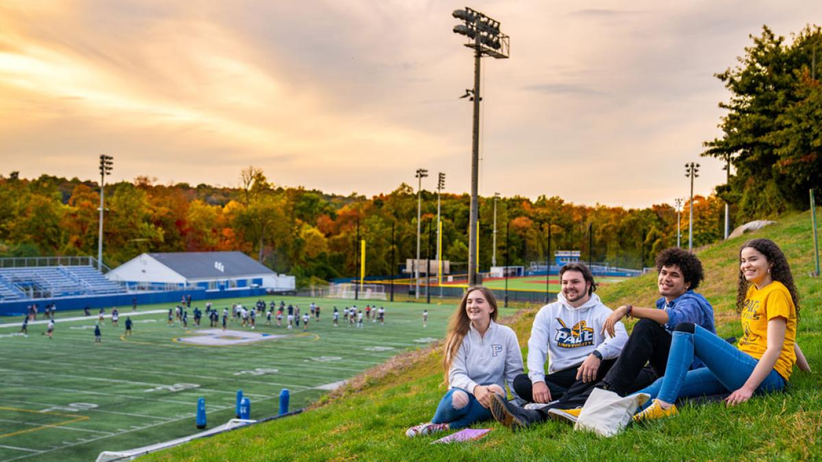 Pace students sit on a grassy hill next to the football field on the Westchester campus