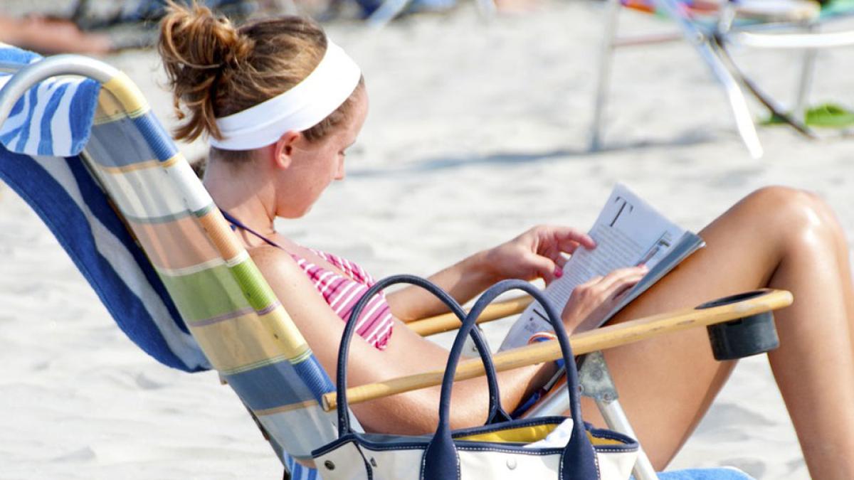 woman in bikini sitting on the beach in a chair reading a magazine