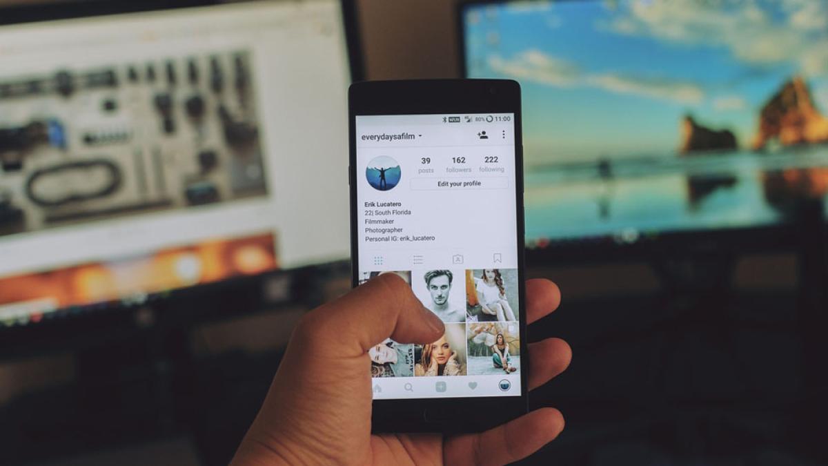 hand holding mobile phone with social media site on screen and two large monitors in background