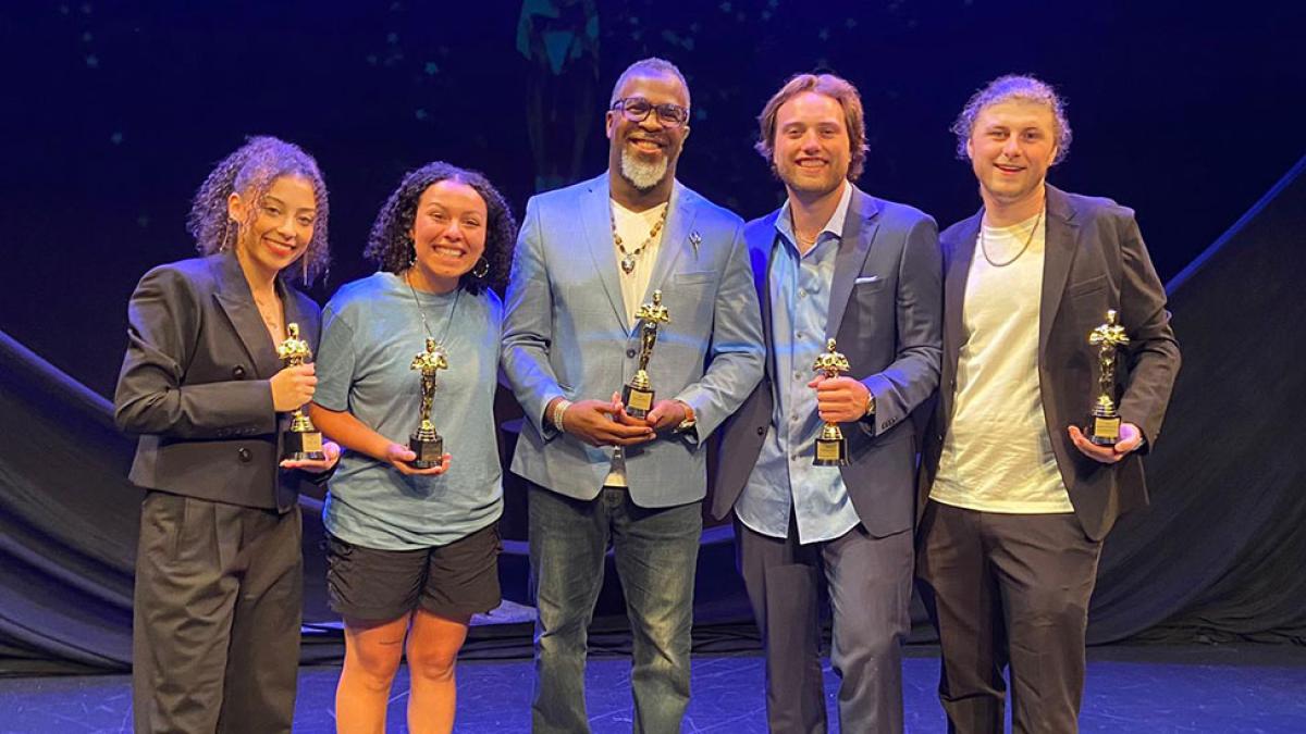 Pace University students Brooke Lyn Sicignano ’25, Ava Resco ’25, Adam Scott ’25, and Gannon Ripchik ’25 stand with Media, Communications, and Visual Arts professor Akil DuPont on stage