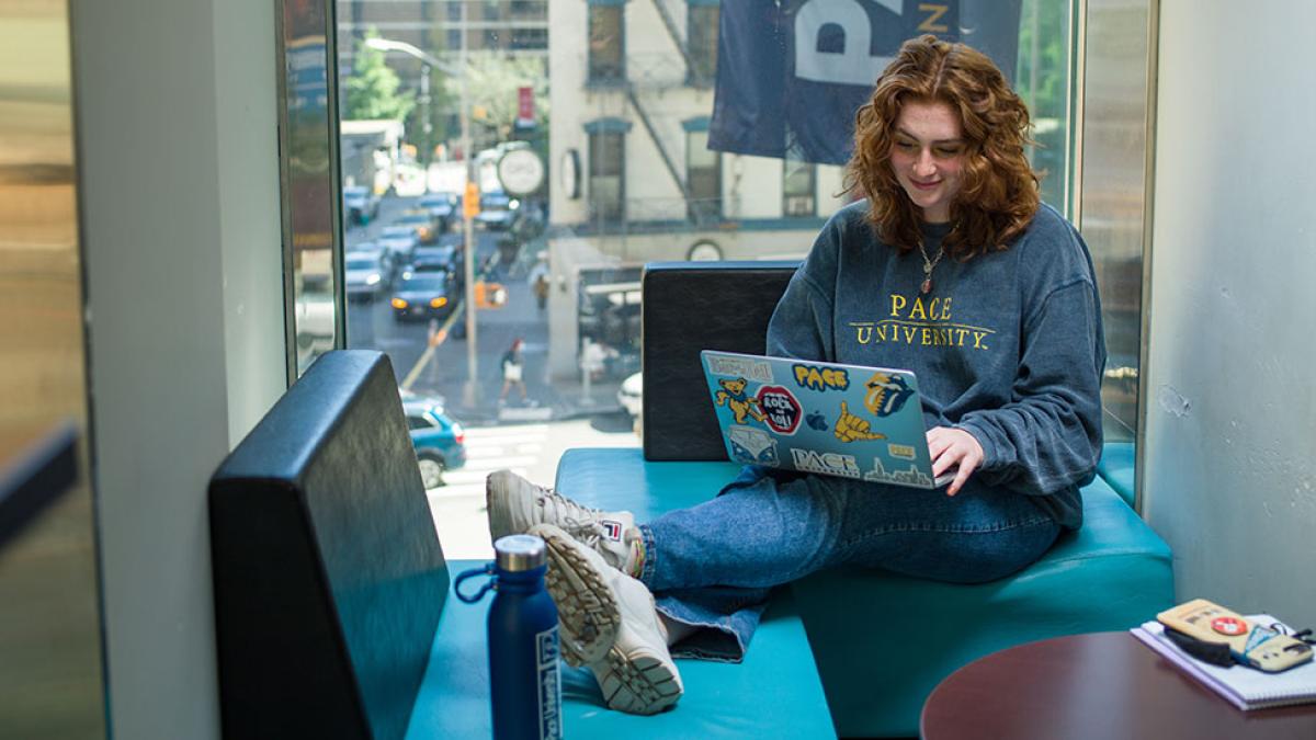 Pace University student sitting near a window looking at her laptop with feet propped up on table