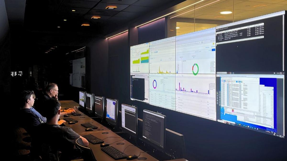 Seidenberg professor Joe Acampora sitting in the Pace Cyber Range with his students, looking at their computers and the large Cyber Range screen which is showing lots of graphs and data.