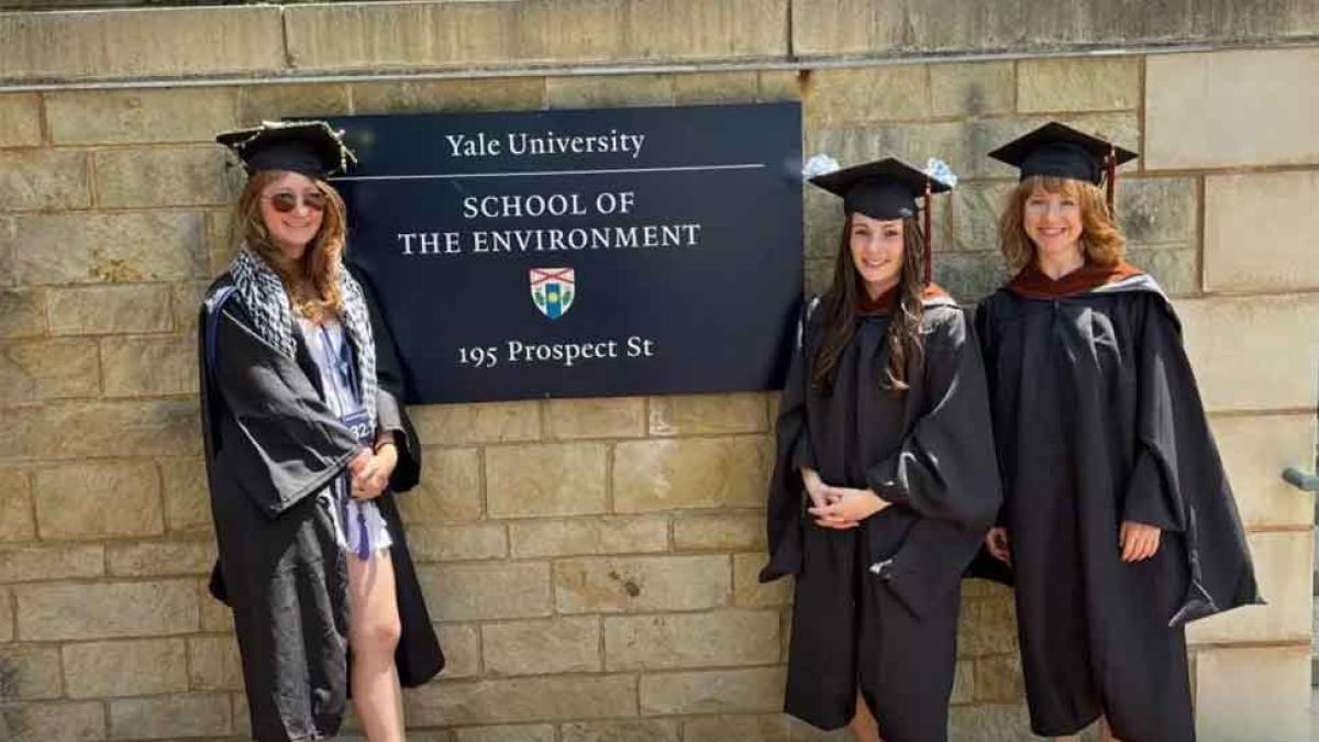 Three Elisabeth Haub School of Law dual degree students in graduate caps and gowns standing outside of Yale Law School sign.