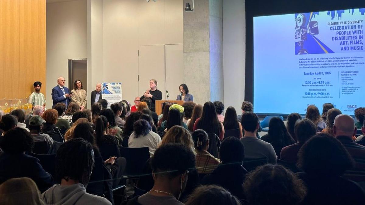 Seidenberg Interim Dean Li-Chiou Chen speaking to a large group of festival attendees in front of a large screen at Pace's Bianco Room for the Disability Film Festival.