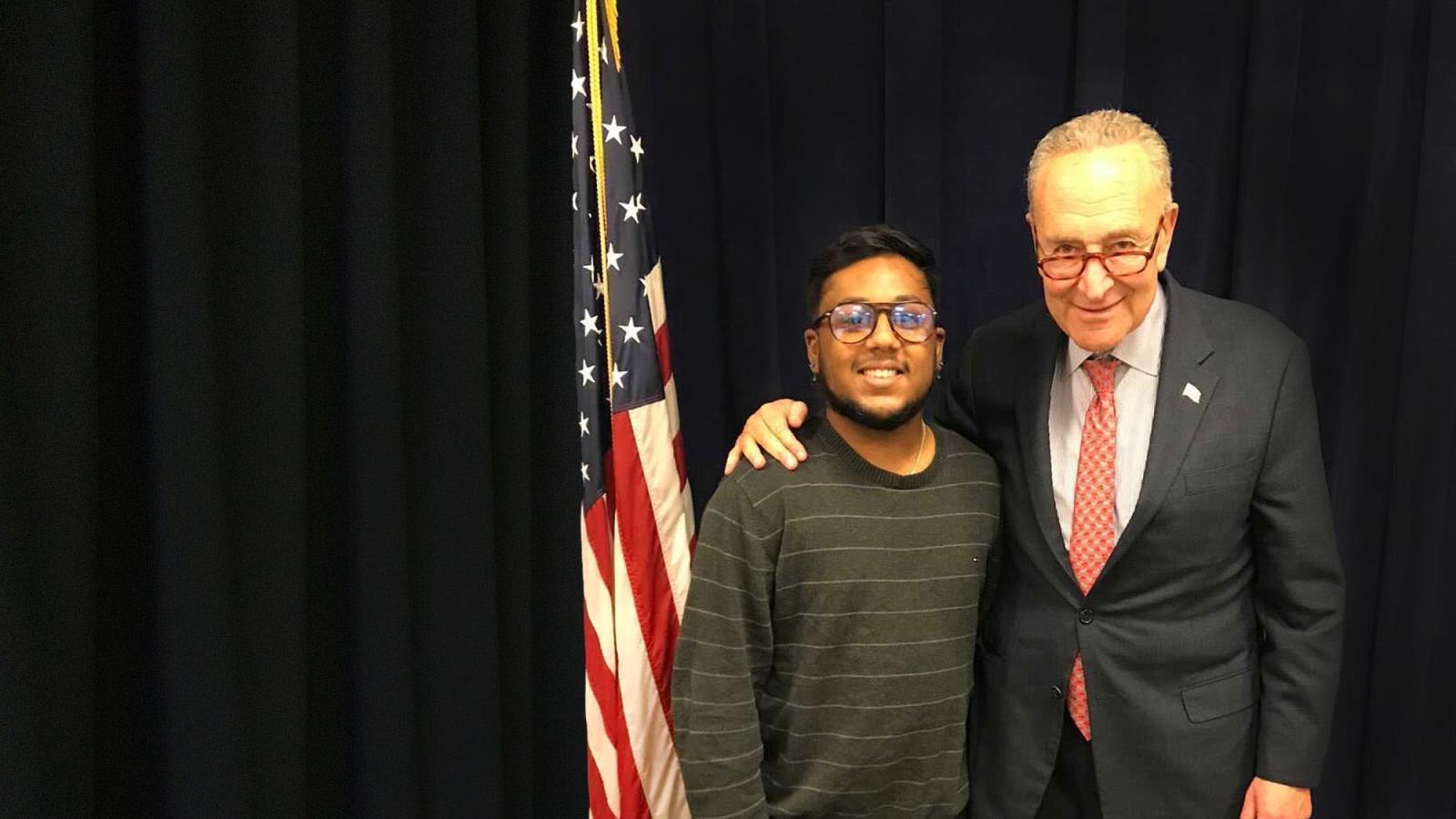 Student Aman Islam stands in front of an American flag with Senate Majority Leader Charles Schumer