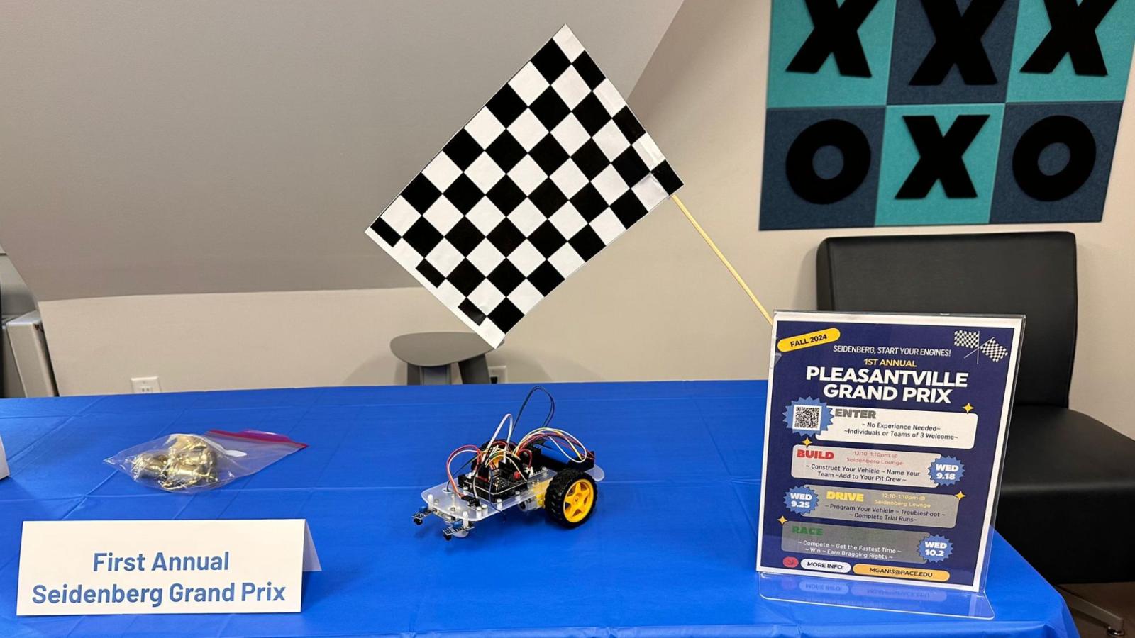 Informational table for the Pleasantville Grand Prix at Pace University, showing a small robot car built by students and a checkered flag.