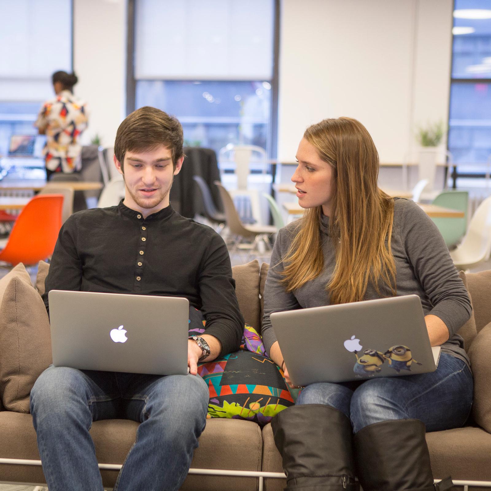 Two students sitting on a couch working on their computers.