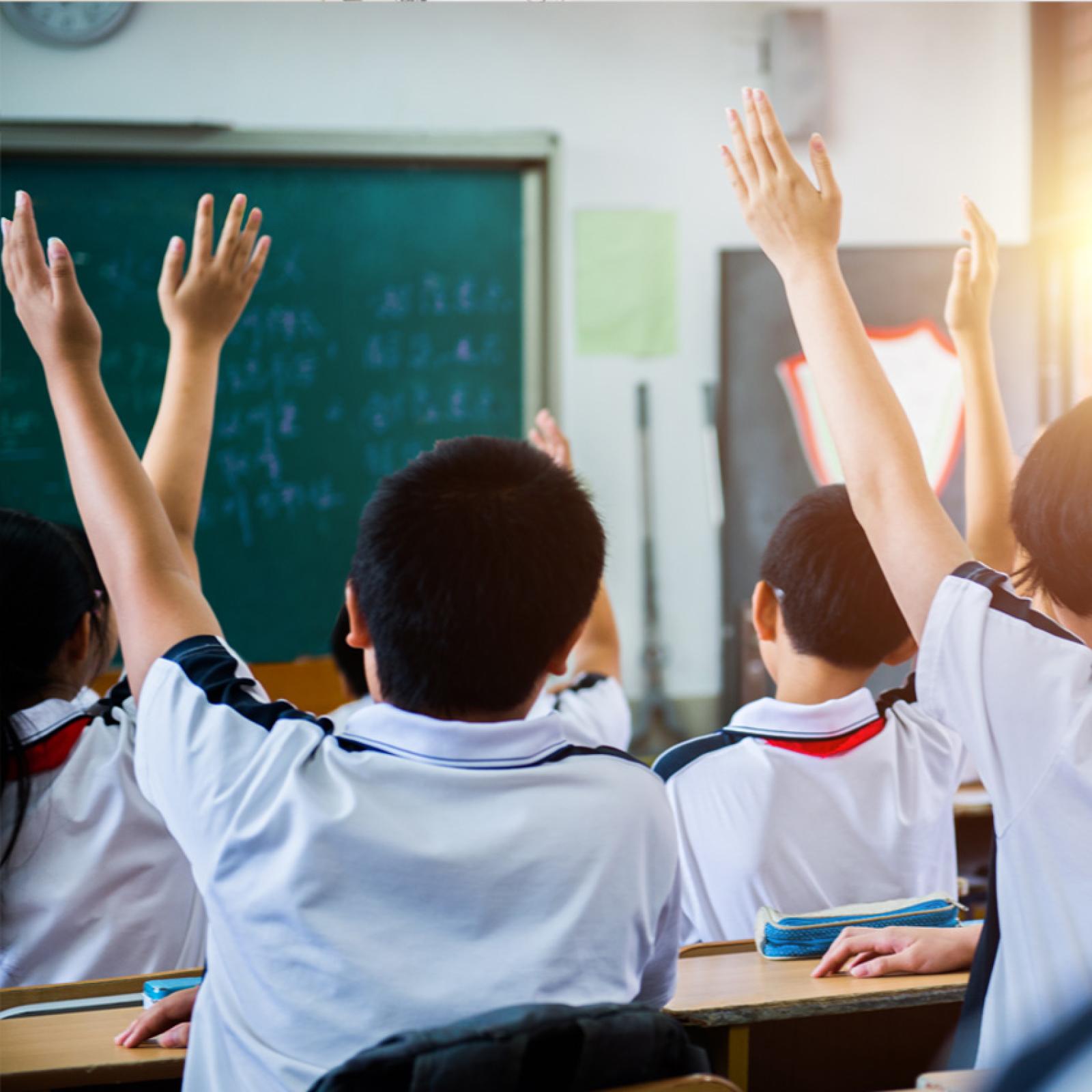 A group of international children in uniform raising their hands in a classroom.