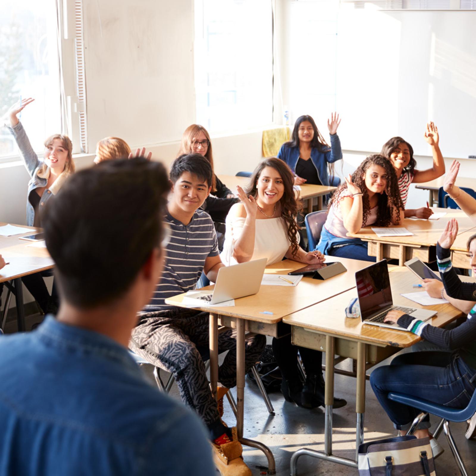 A teacher standing in front of a high school classroom where students are raising their hands.