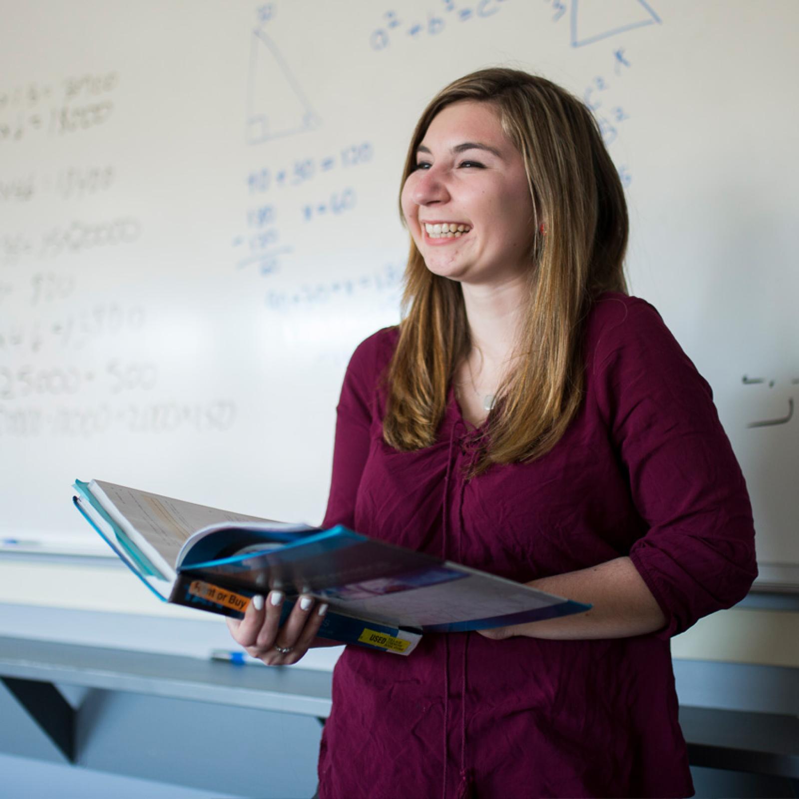 A teacher in front of a whiteboard smiling while holding a textbook.