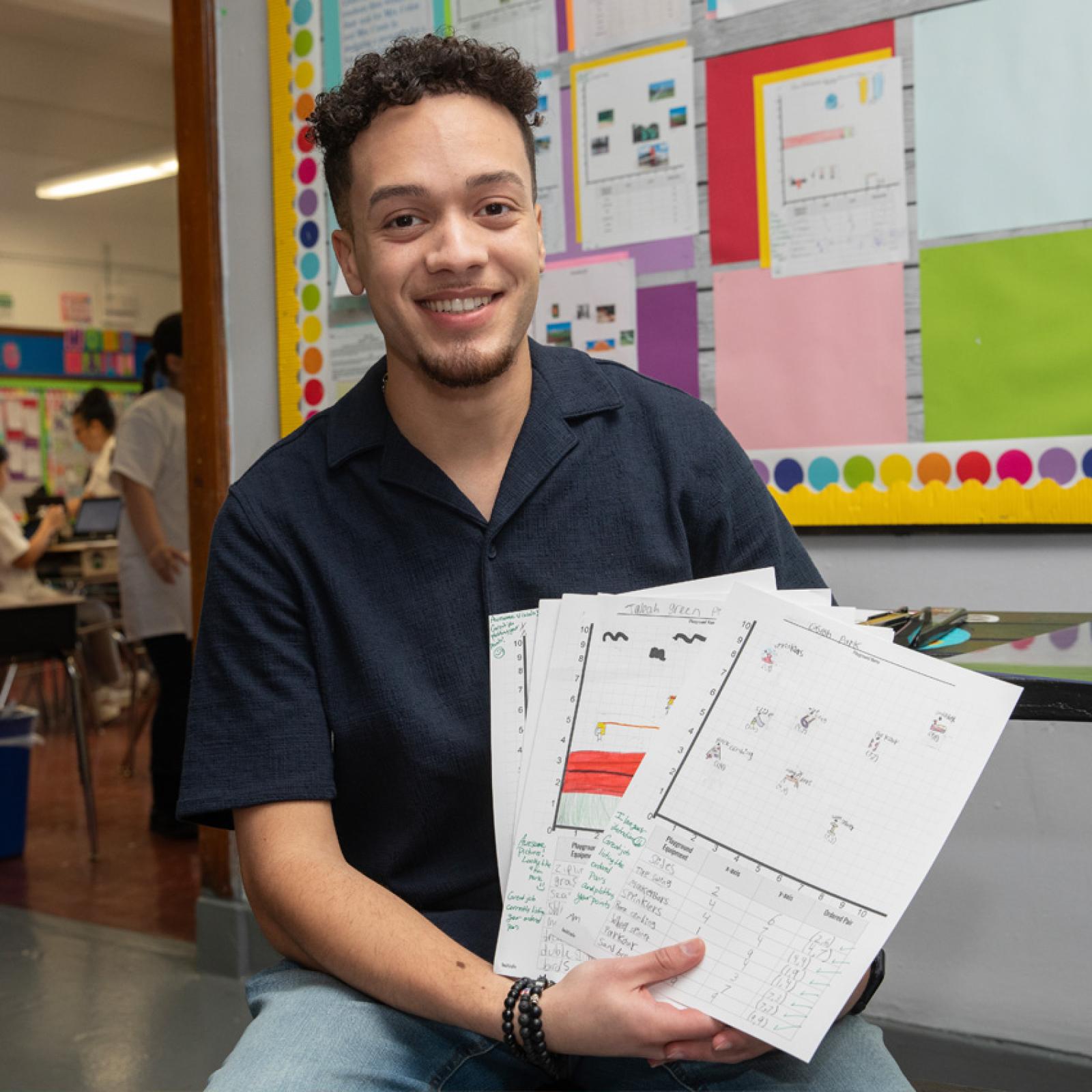 A male teacher in the hallway of an elementary school holding worksheets.