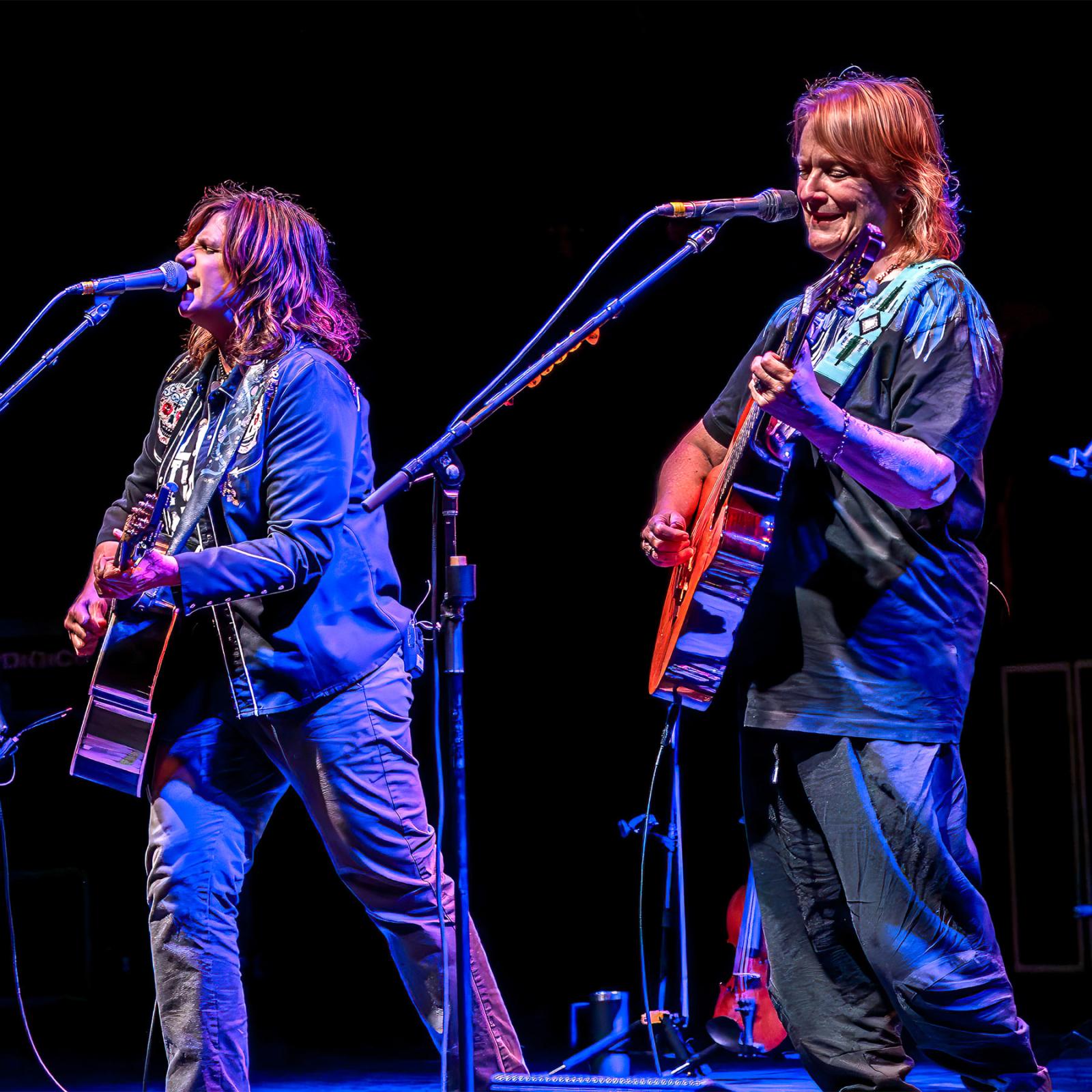 Indigo Girls band members Amy Ray and Emily Saliers play guitars and sing into microphones joined on-stage by their violinist Lyris Hung.