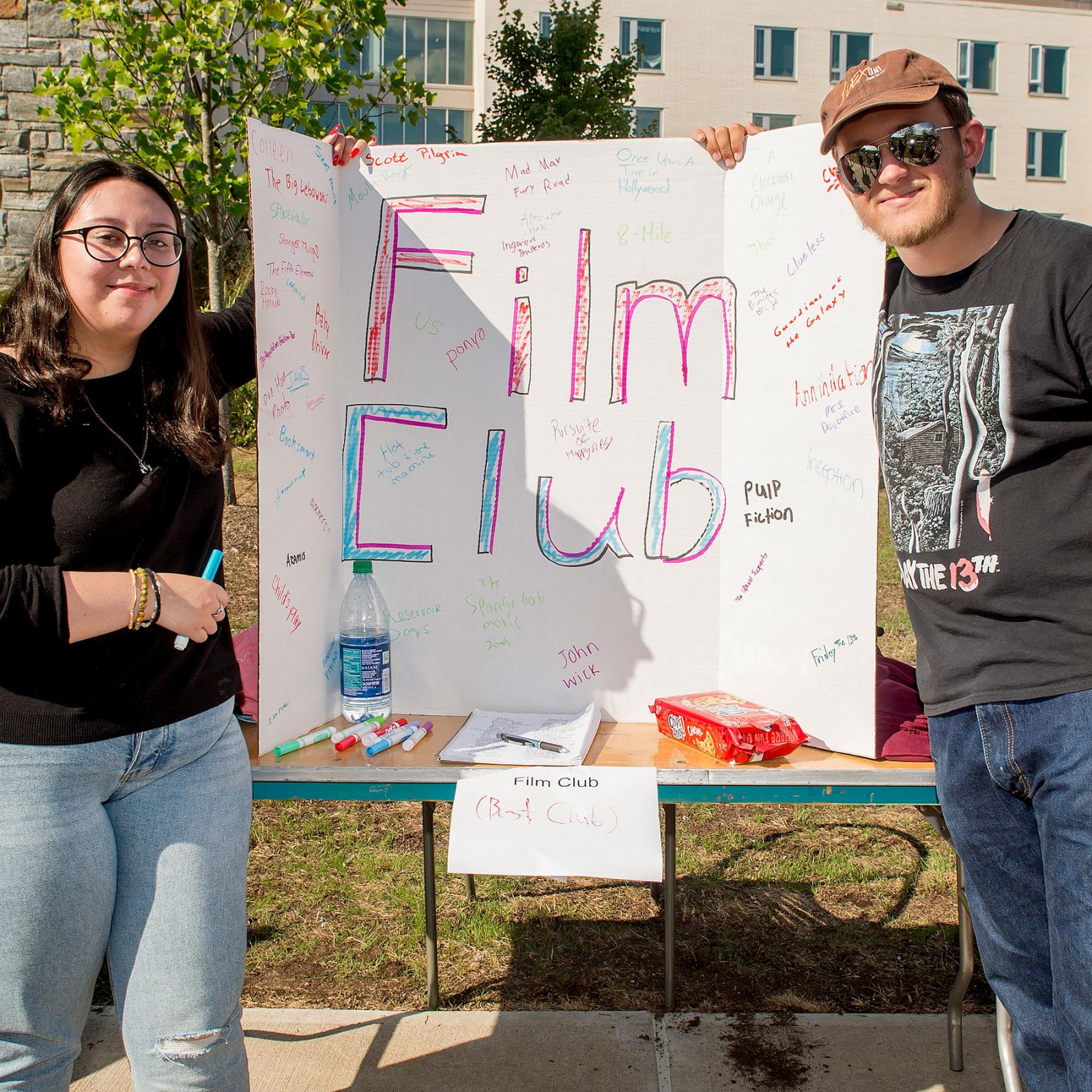 Two Pace University Film Club members holding a large Film Club sign on a table outside